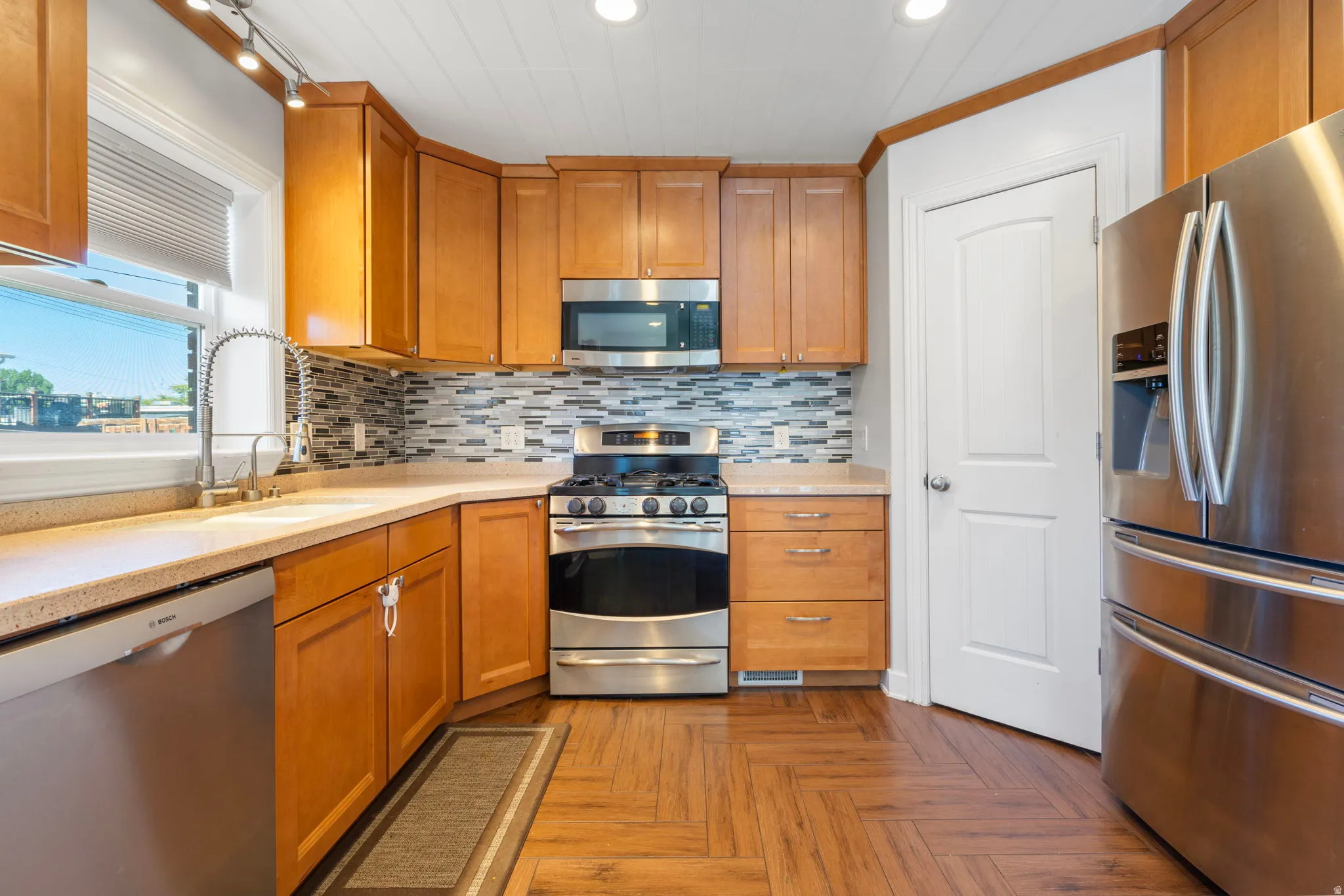 Kitchen with stainless steel appliances, wood finish cabinetry, parquet floors, decorative backsplash, and light stone counters