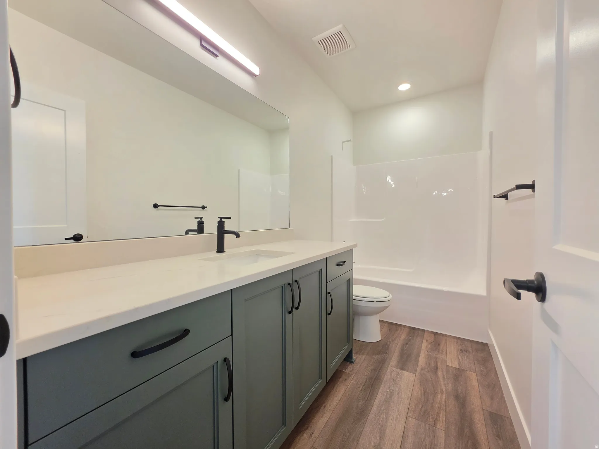 Bathroom featuring a green vanity with a light countertop, wood-finish flooring, a full-width mirror, and a white tub with shower surround