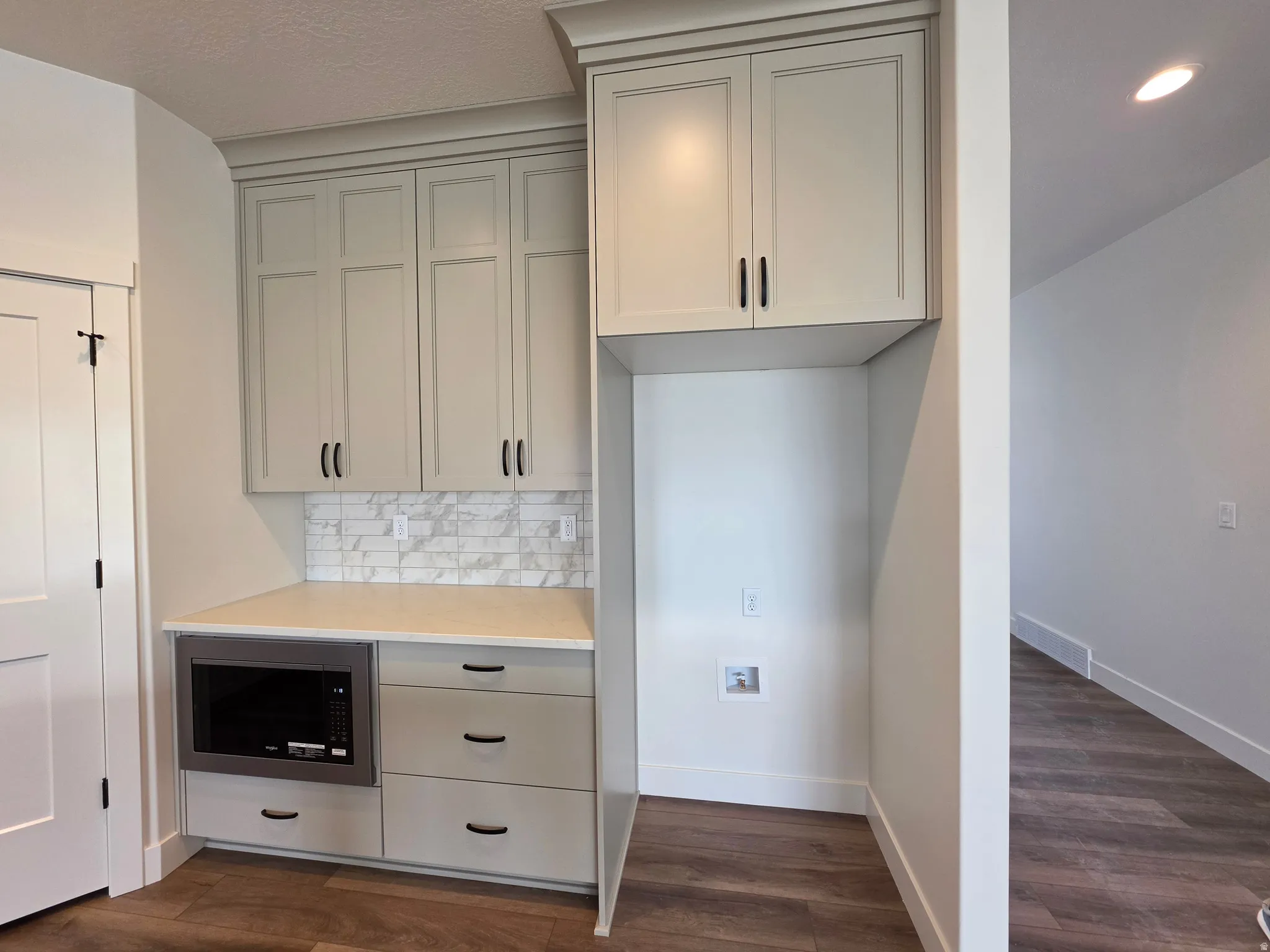 Built-in microwave with cabinet surround, light-toned countertops, subway tile backsplash, shaker-style cabinetry with dark hardware, and wood-finish flooring