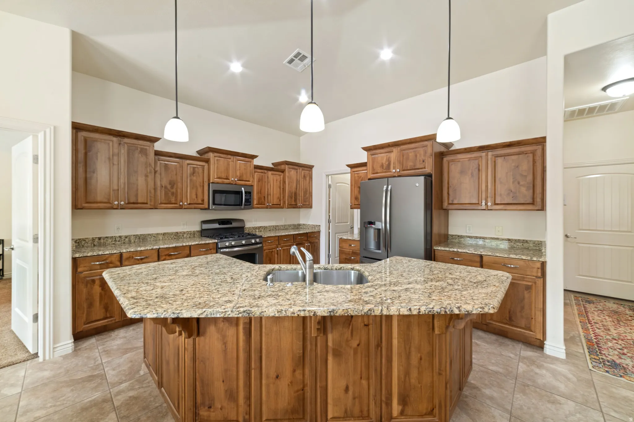 Kitchen featuring wood finish cabinetry, stainless steel appliances, light stone countertops, an island with sink, and pendant lighting