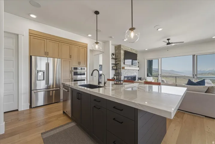 Dual tone kitchen with open floor plan, stainless steel appliances, a stone fireplace, hanging light fixtures, and a center island with sink