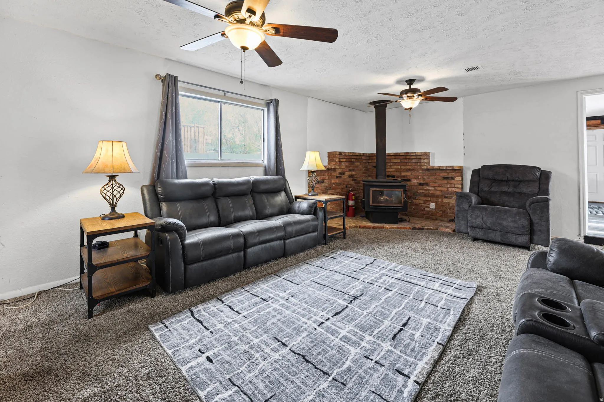 Living room with a wood stove, carpet flooring, a ceiling fan, and a textured ceiling