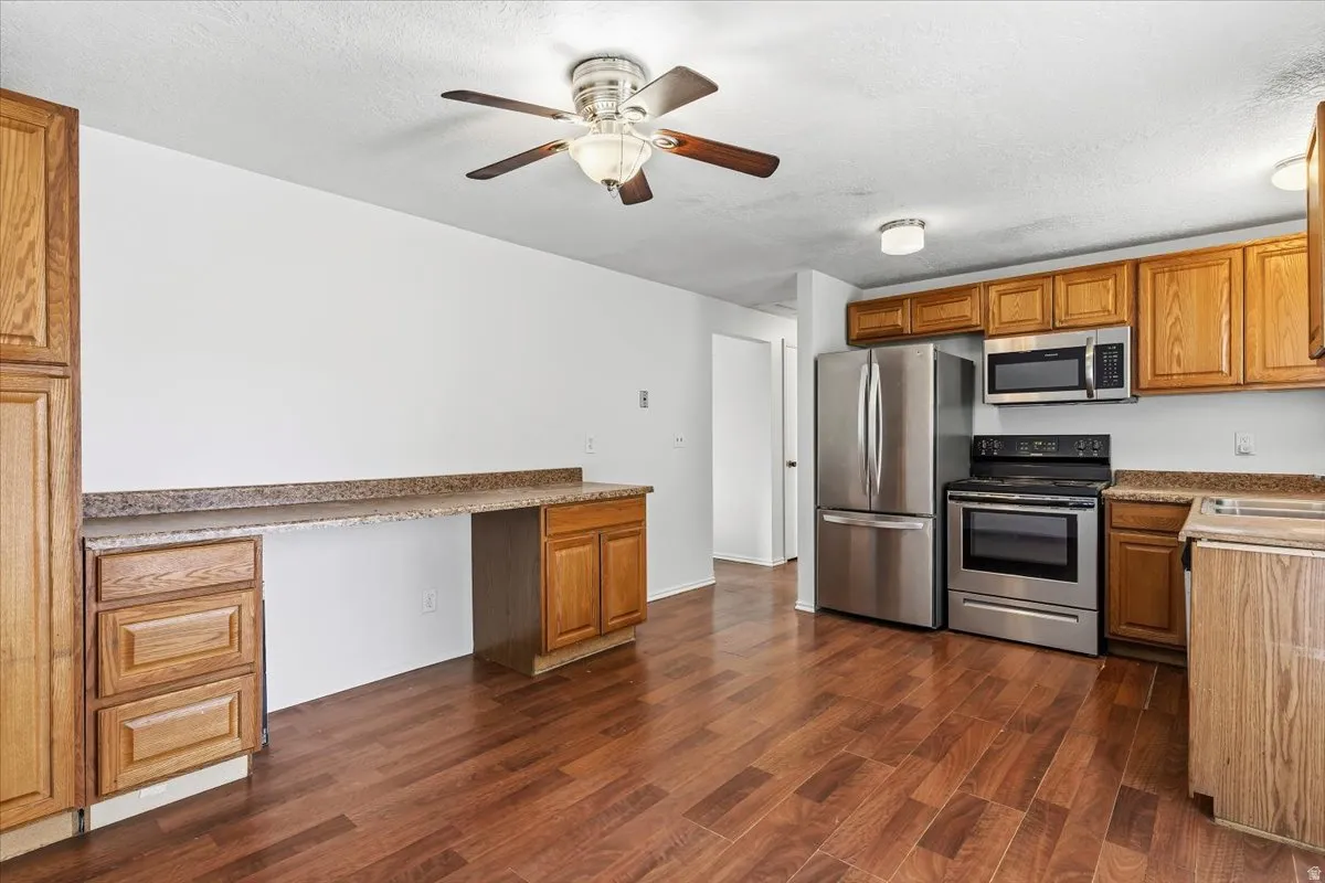 Kitchen featuring stainless steel appliances, built in study area, ceiling fan, wood finish cabinetry, and dark wood-style floors
