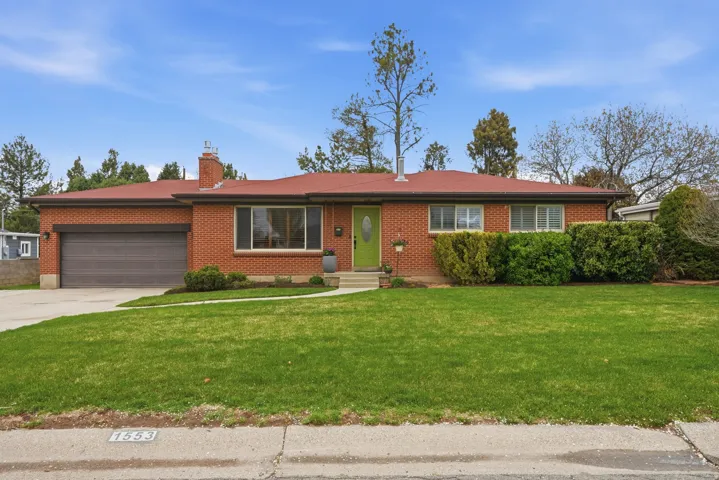 Rambler-style home featuring a garage, a front lawn, driveway, brick siding, and a chimney