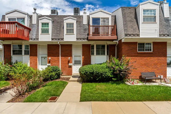 Brick and siding exterior featuring multiple dormer windows, a private balcony with wood railing, a paved walkway, and a manicured lawn with established landscaping
