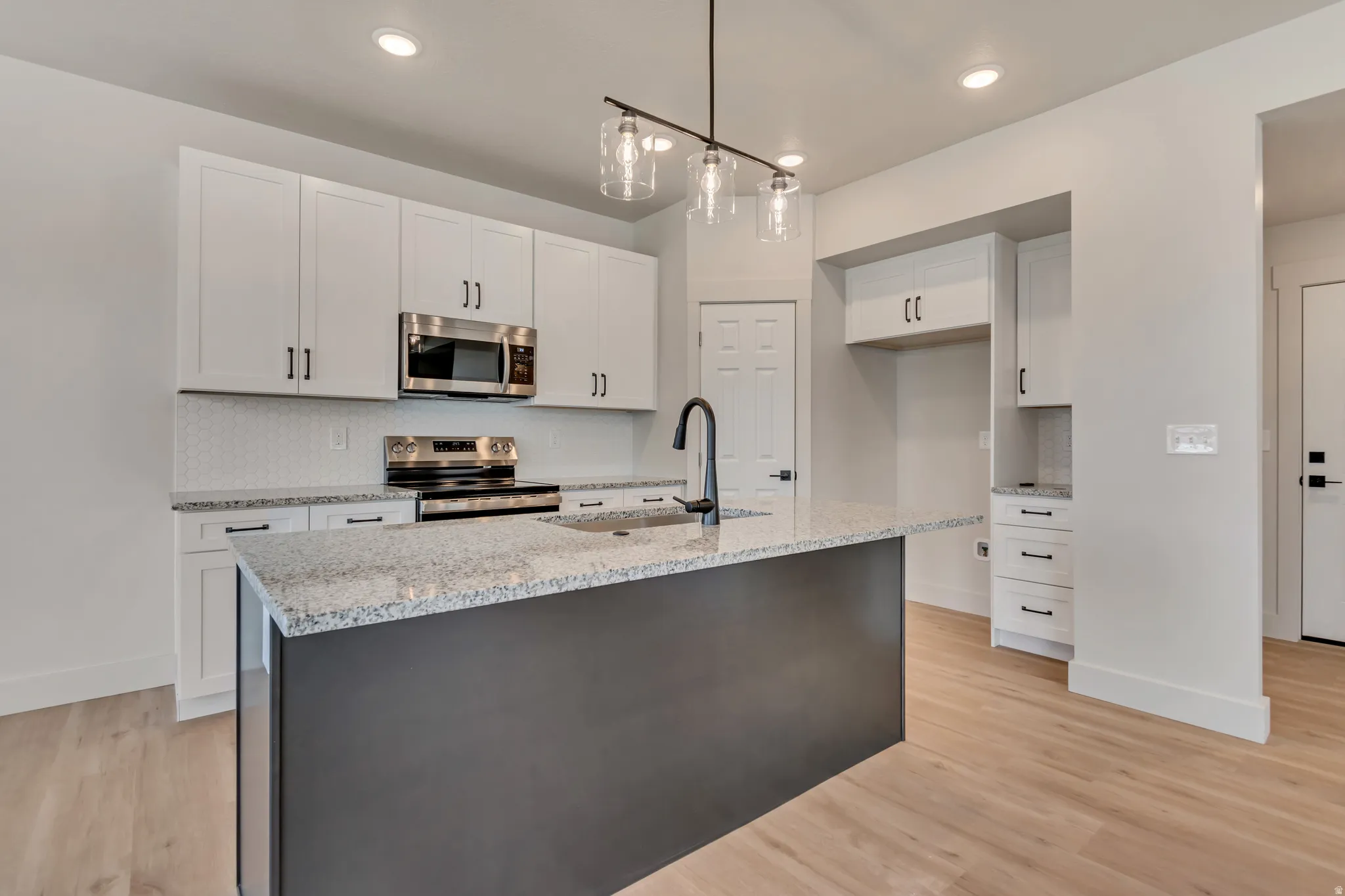 Two tone kitchen with stainless steel appliances, light wood finished floors, light stone counters, hanging light fixtures, and an island with sink