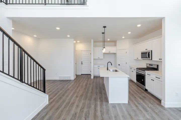 Open concept living area featuring wood-finish flooring, a kitchen island with sink, white shaker cabinetry, stainless steel appliances, and a black wrought iron staircase railing