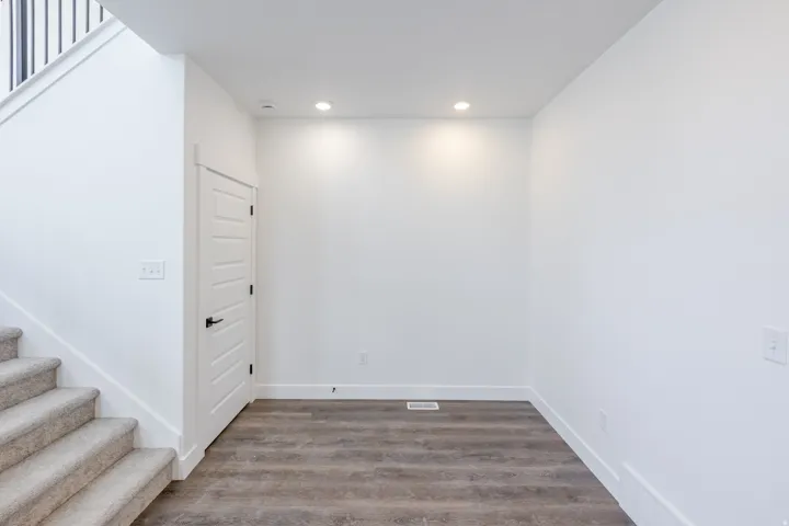 Recessed ceiling lighting, wood-finish flooring, white paneled interior door with matte black hardware, white walls, and carpeted stairs with black metal balusters