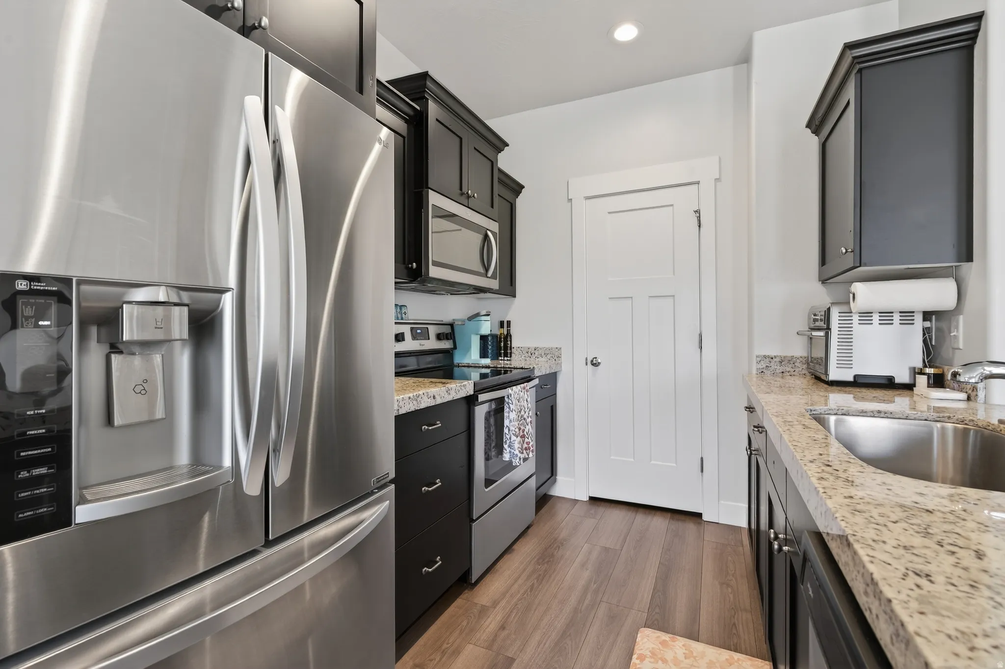 Kitchen with stainless steel appliances, dark cabinetry, light stone counters, dark wood-style floors, and recessed lighting