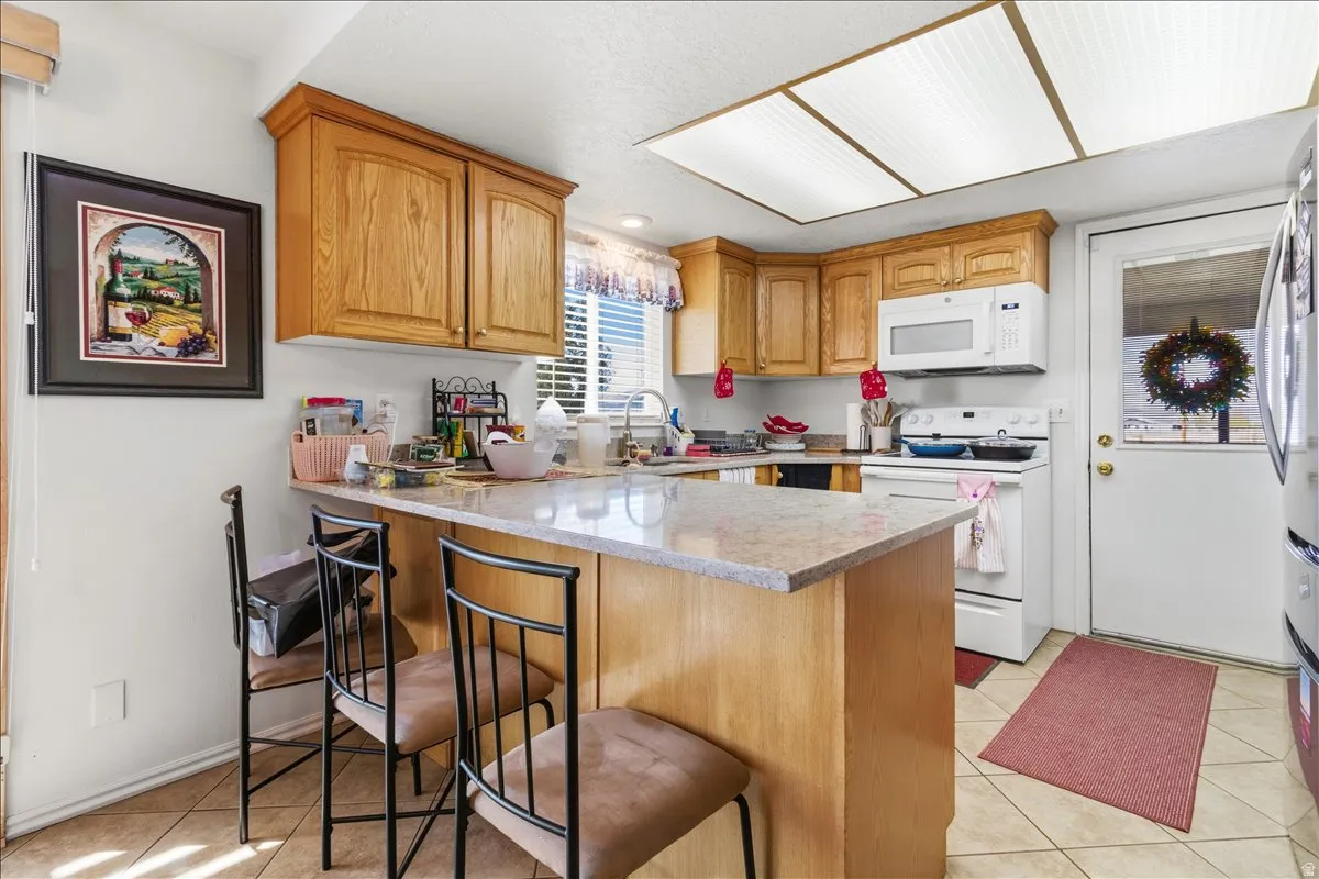 Kitchen featuring white appliances, a peninsula, light tile patterned floors, a breakfast bar, and light stone countertops
