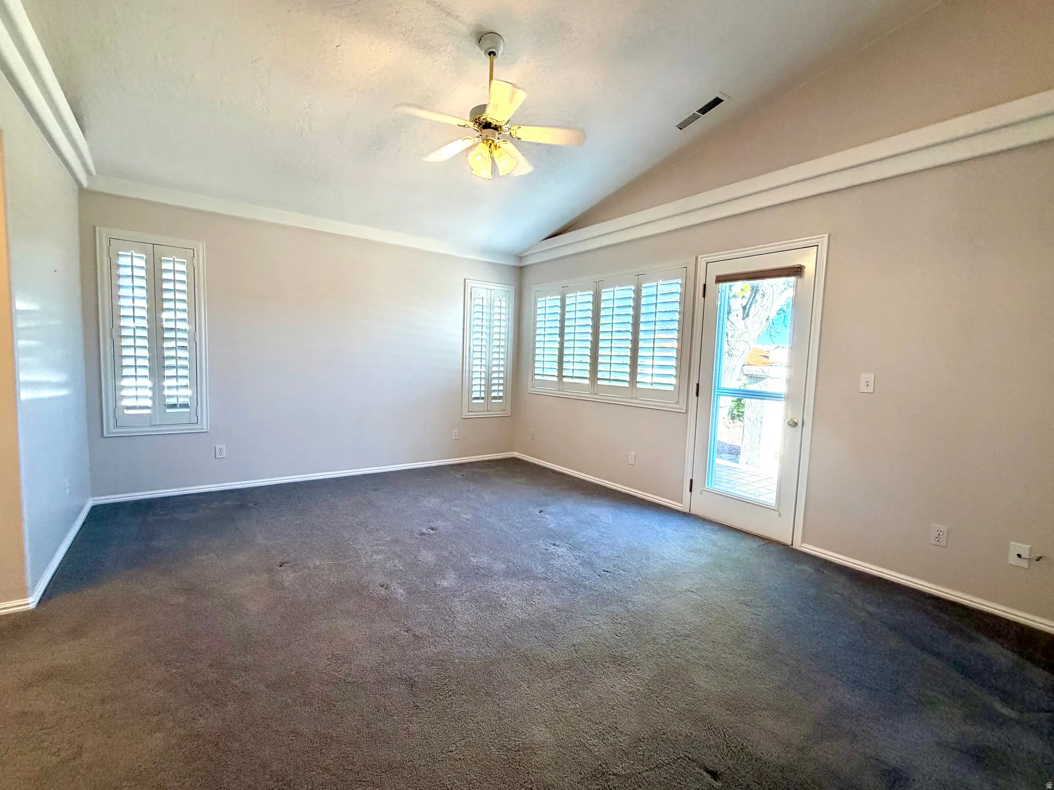 Spacious interior room featuring a vaulted ceiling with a ceiling fan, plush gray carpeting, white plantation shutters on multiple windows, and a glass-paneled door