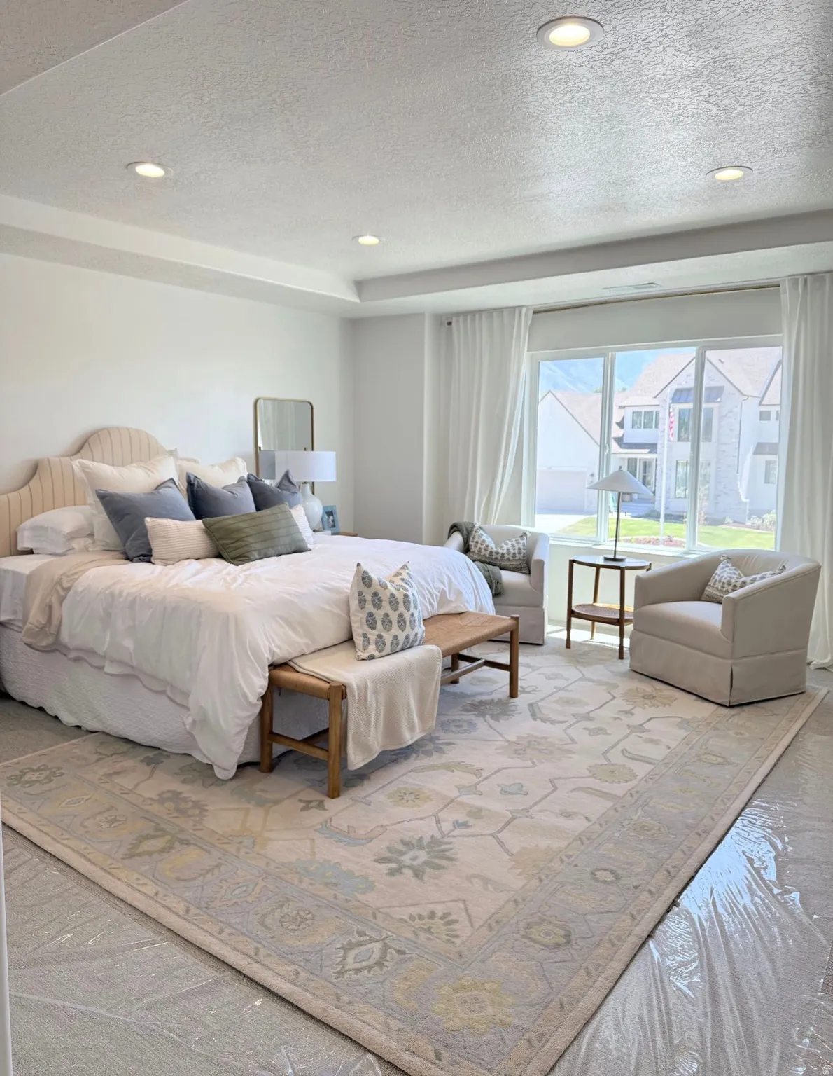 Recessed lighting illuminates a spacious room featuring a tray ceiling and a large window with white drapes
