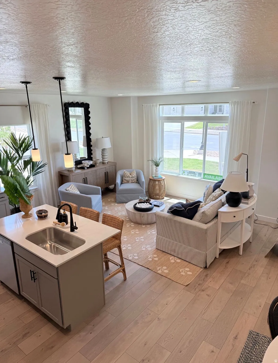 Open-concept living area featuring light wood-finish flooring, a kitchen island with a built-in sink and seating, and a large window providing natural light