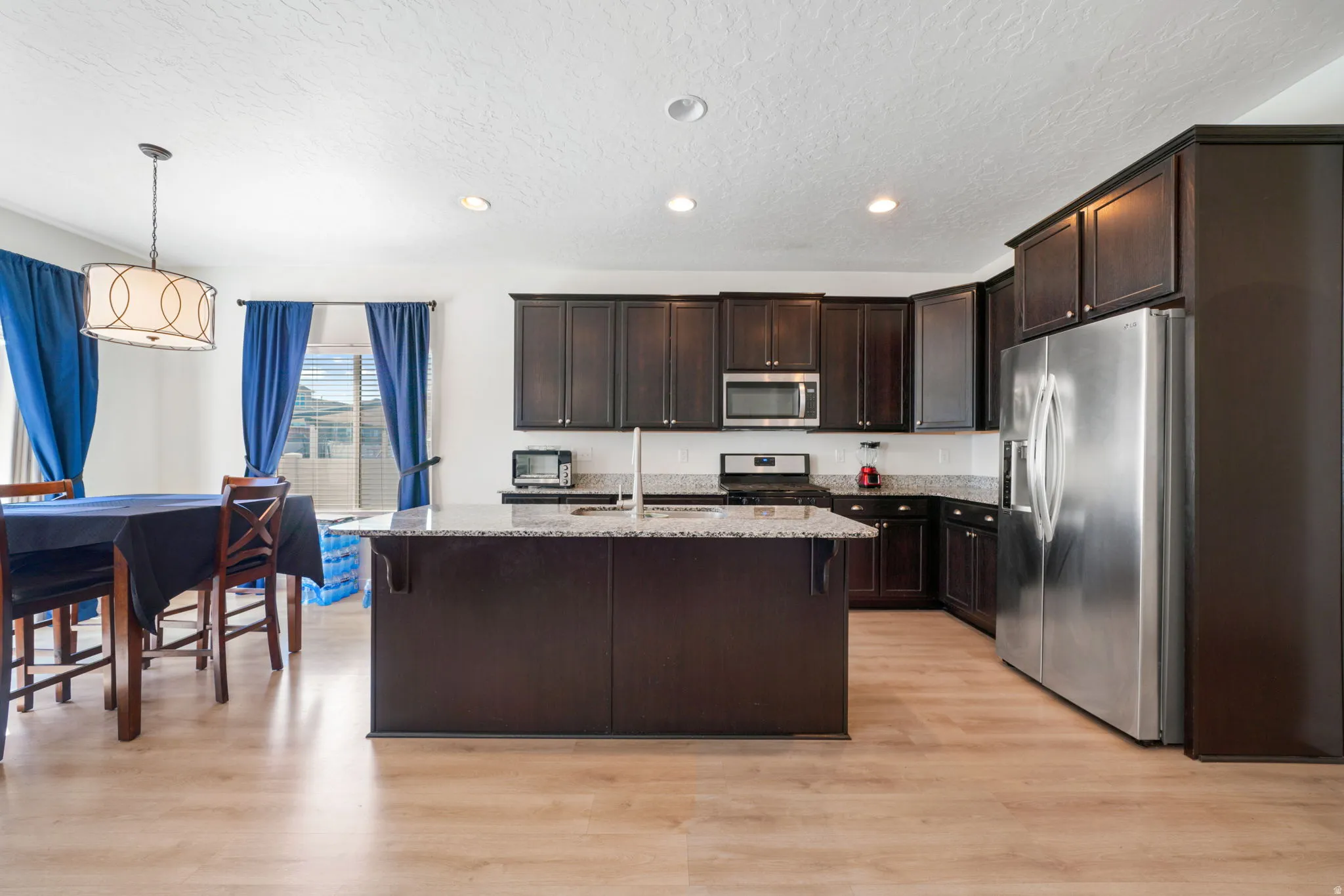 Kitchen with stainless steel appliances, dark wood finish cabinetry, light stone countertops, light wood-style floors, and an island with sink