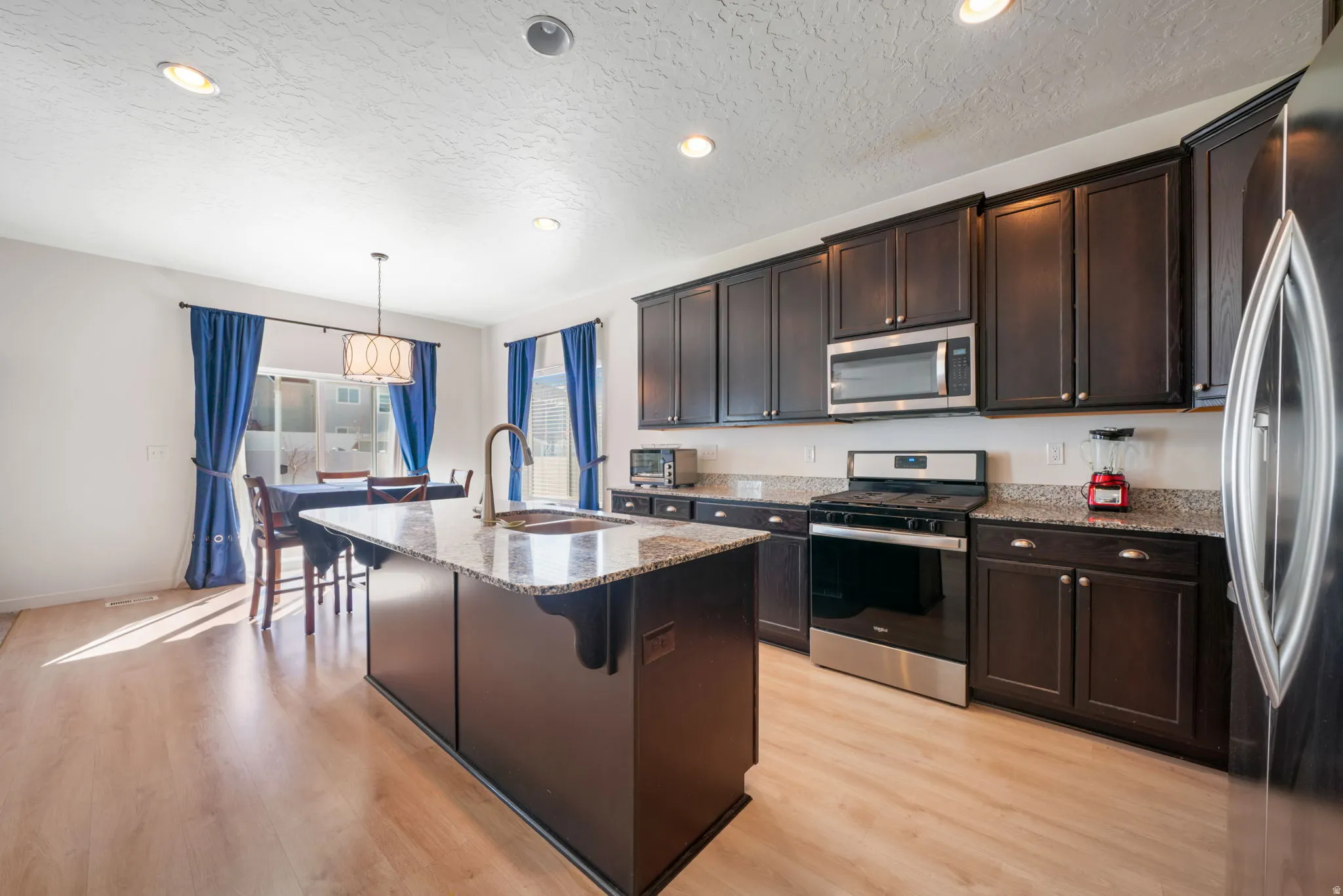 Kitchen with light stone countertops, stainless steel appliances, dark wood finish cabinetry, a center island with sink, and light wood-style floors