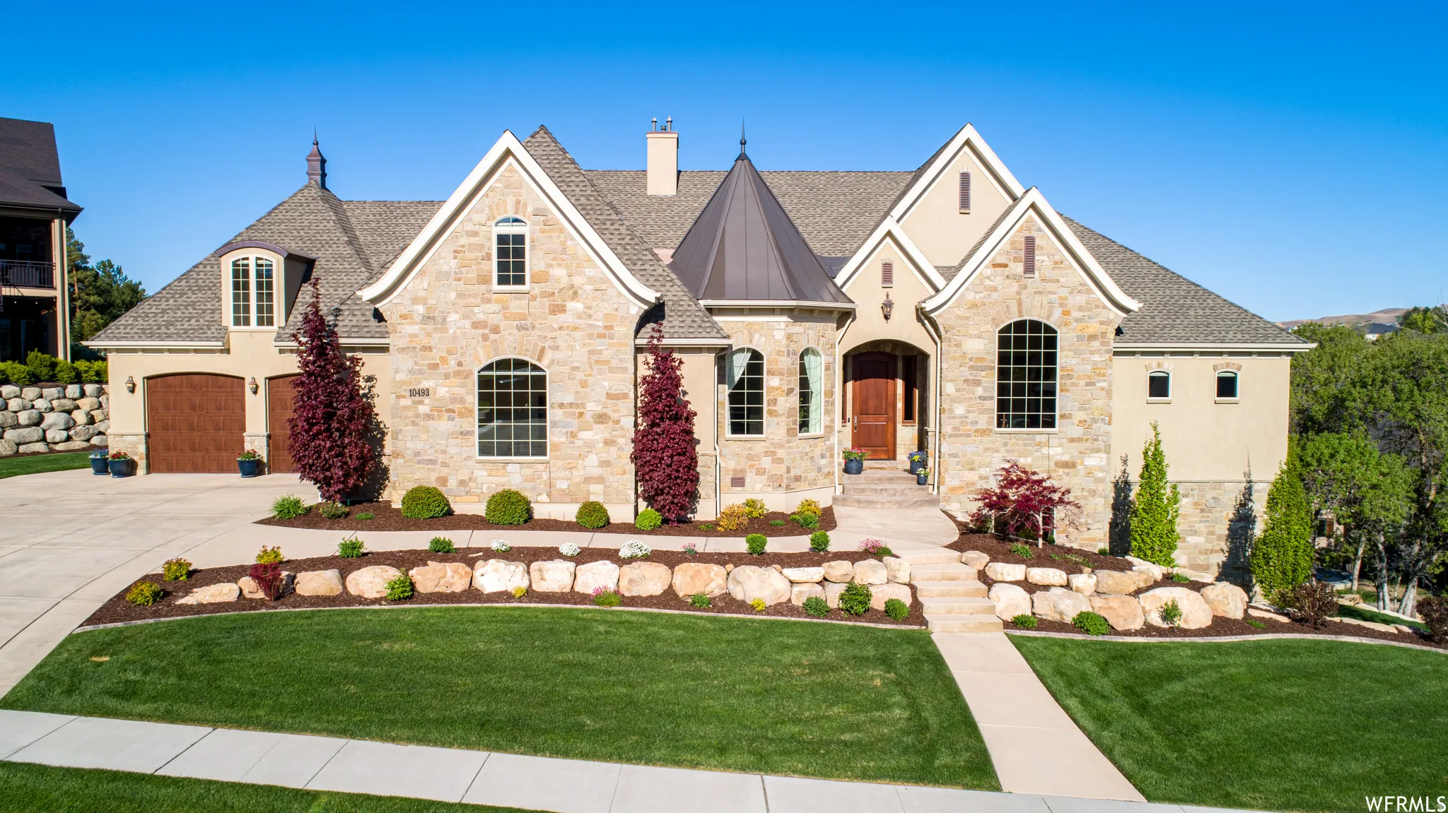 Stone and stucco exterior featuring a prominent turret, arched windows, a wood-finish front door, a concrete driveway with wood-finish garage doors, and landscaped beds with rock accents