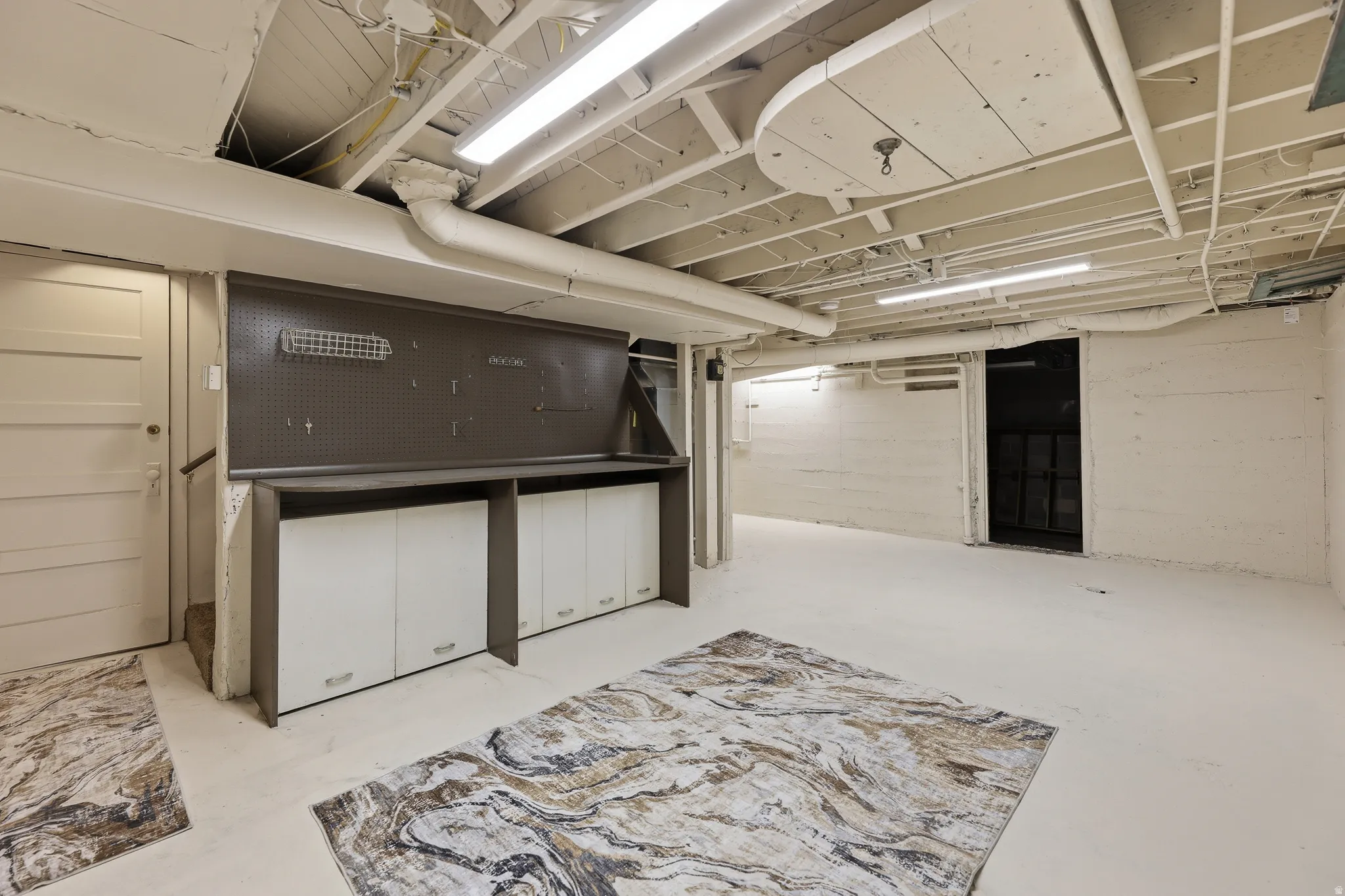 Spacious basement area featuring a built-in workbench with integrated storage cabinetry and pegboard, exposed ceiling joists, white-painted walls, and fluorescent lighting fixtures