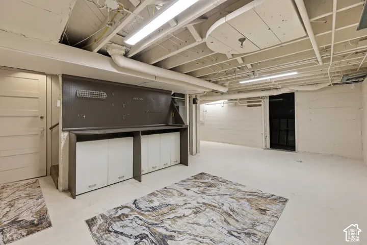 Spacious basement area featuring a built-in workbench with integrated storage cabinetry and pegboard, exposed ceiling joists, white-painted walls, and fluorescent lighting fixtures