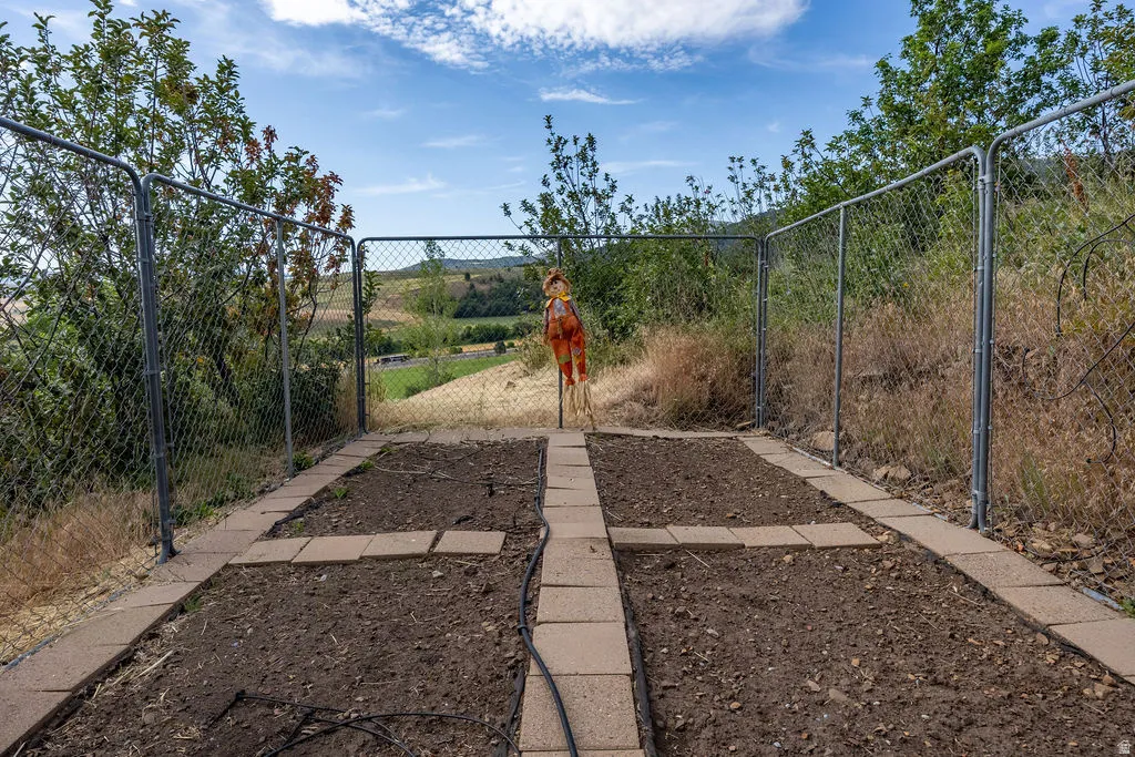 Raised garden bed with paver stone borders and central path, enclosed by a chain-link fence, featuring a garden hose