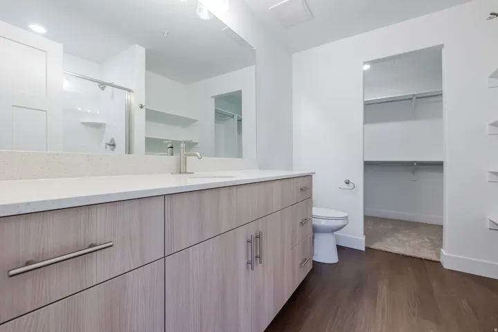 Wood-finish flooring, light wood-look vanity with metal hardware, light-colored countertop, and a built-in shower with clear glass enclosure
