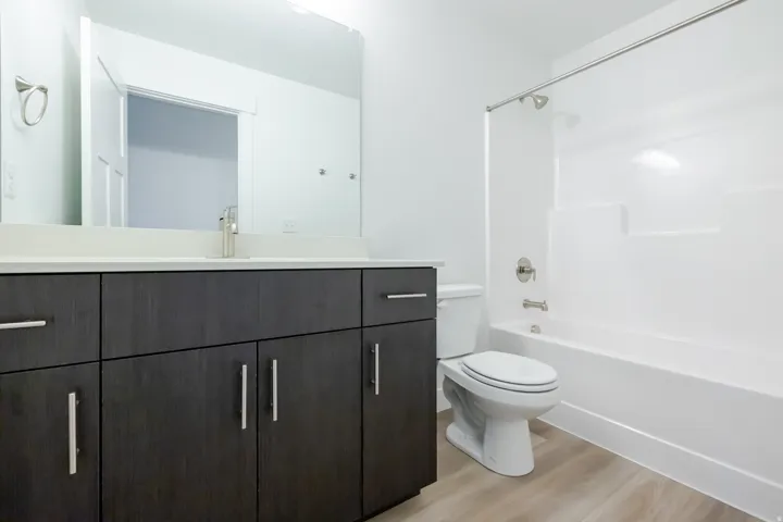 Bathroom featuring a dark wood-finish vanity with a light-toned countertop, a large mirror, a white toilet, and a white bathtub with a shower
