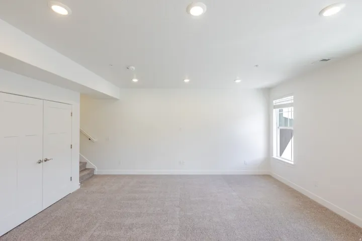 Light-colored neutral carpeting, recessed ceiling lighting, a single pane window with horizontal blinds, and white bi-fold doors