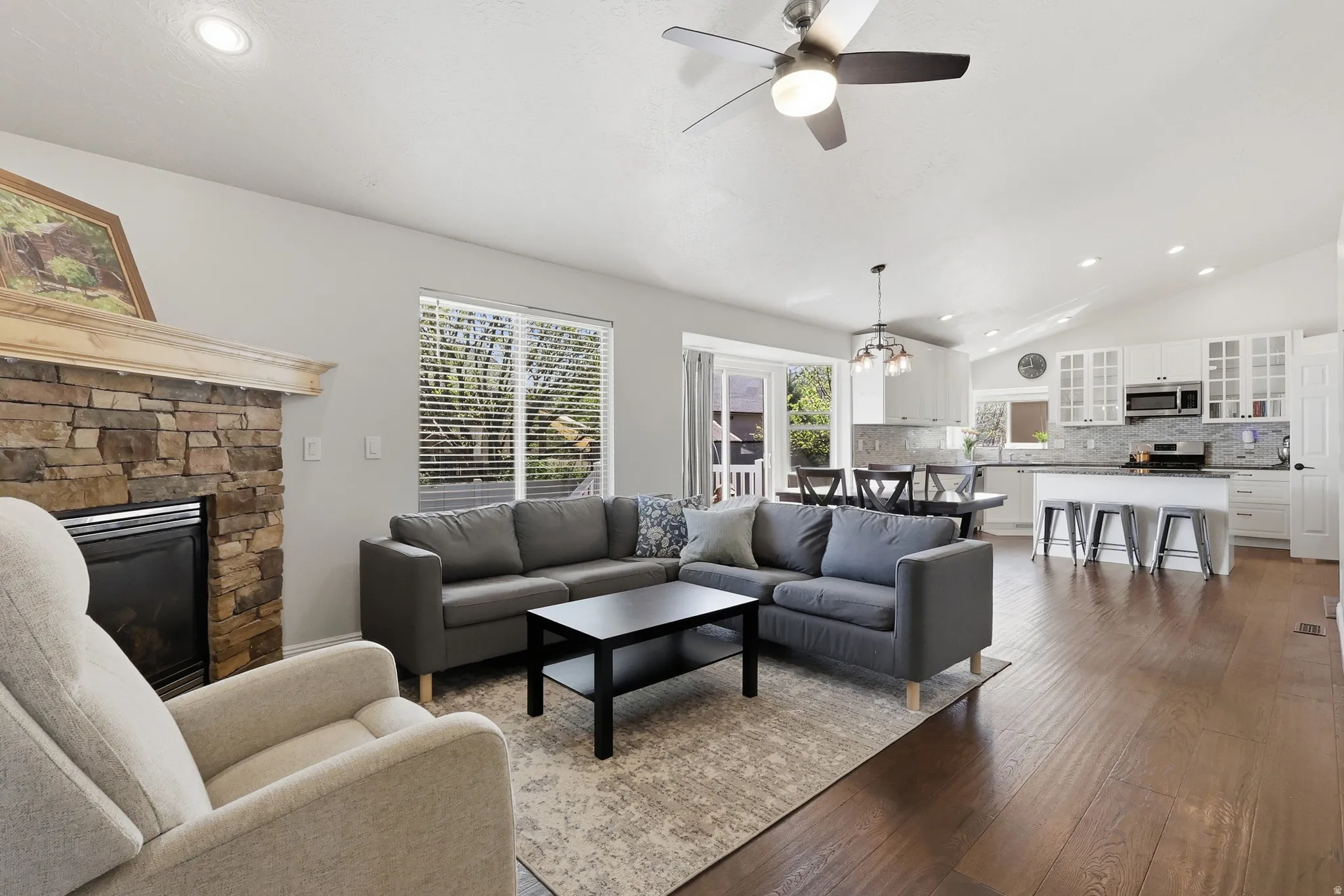 Living room with vaulted ceiling, plenty of natural light, a fireplace, wood flooring, dining table & doors to the back yard