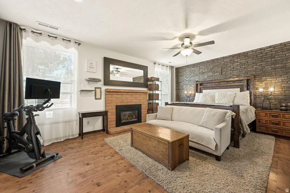 Bedroom featuring brick wall, wood-type flooring, a brick fireplace, a textured ceiling, and ceiling fan