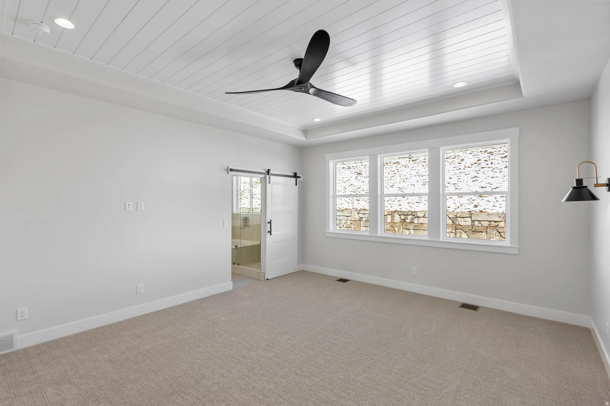 Unfurnished bedroom featuring light carpet, a wood tray ceiling, a barn door, ensuite bathroom, and a ceiling fan