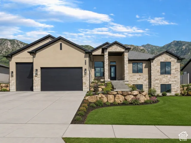 Stucco and stone exterior featuring a multi-gabled roofline, three-car garage, concrete driveway, and terraced landscaping with stacked stone retaining walls against a mountain backdrop