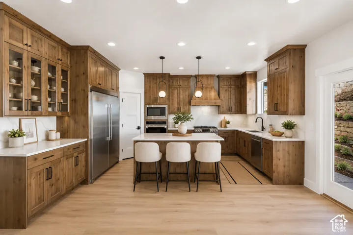 Spacious kitchen featuring warm wood cabinetry, stainless steel appliances, a generous central island with seating, white countertops, and wood-finish flooring
