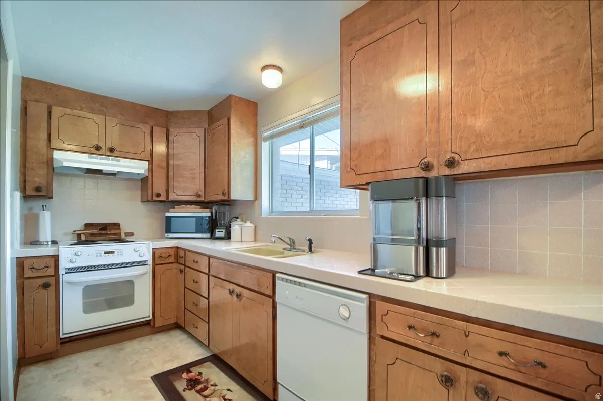 Kitchen featuring white appliances, decorative backsplash, wood finish cabinetry, light flooring, and tile countertops