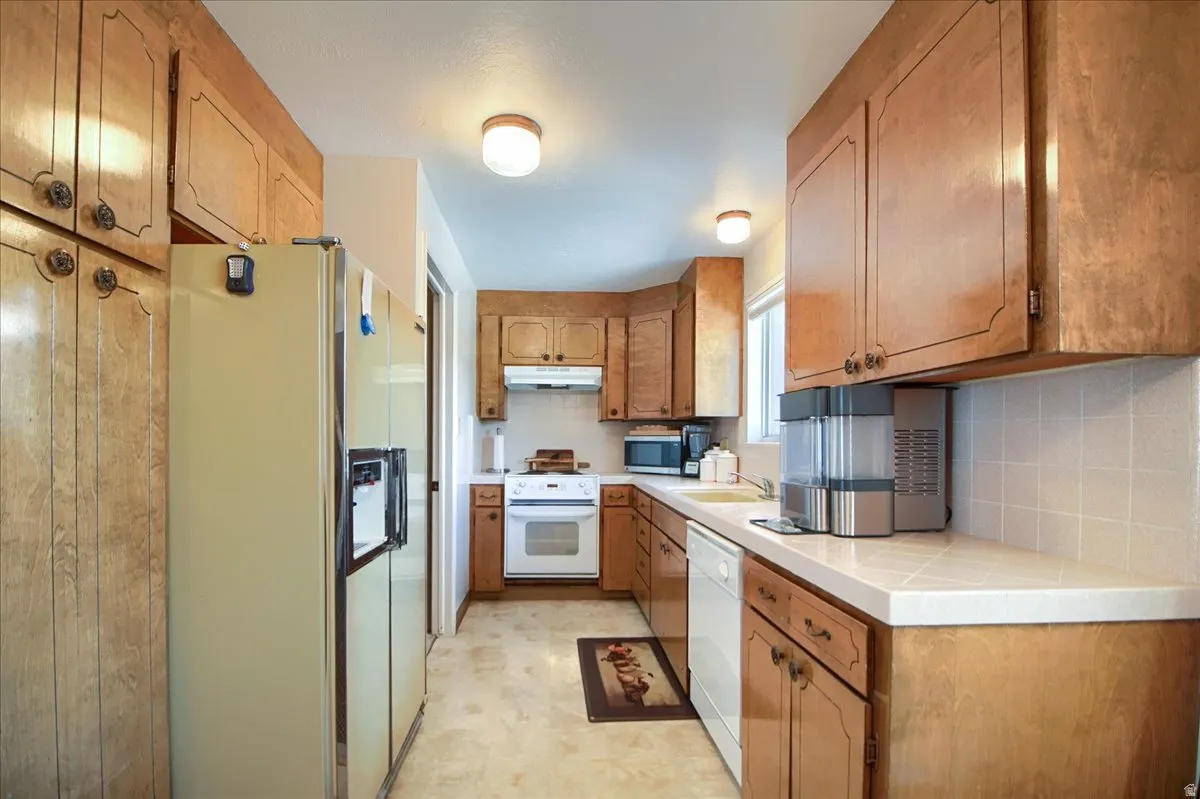 Kitchen with white appliances, light flooring, wood finish cabinetry, tile counters, and tasteful backsplash
