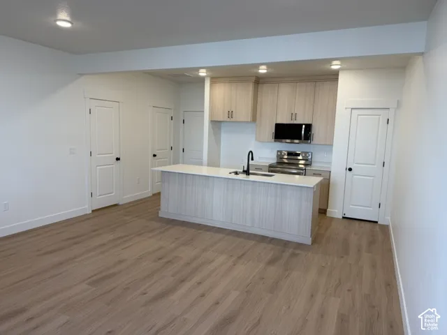 Kitchen featuring stainless steel appliances, light wood finished floors, an island with sink, light wood finish cabinetry, and light stone countertops