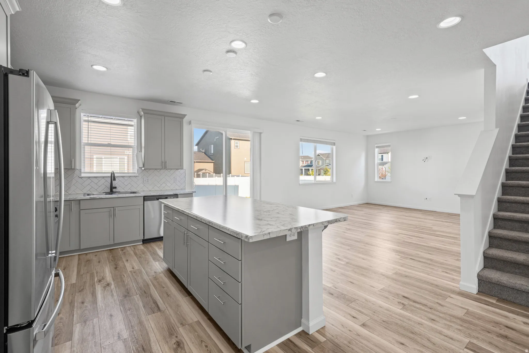 Open-concept living space featuring a kitchen island with stone countertop, stainless steel appliances, wood-finish flooring, recessed lighting, and a carpeted staircase