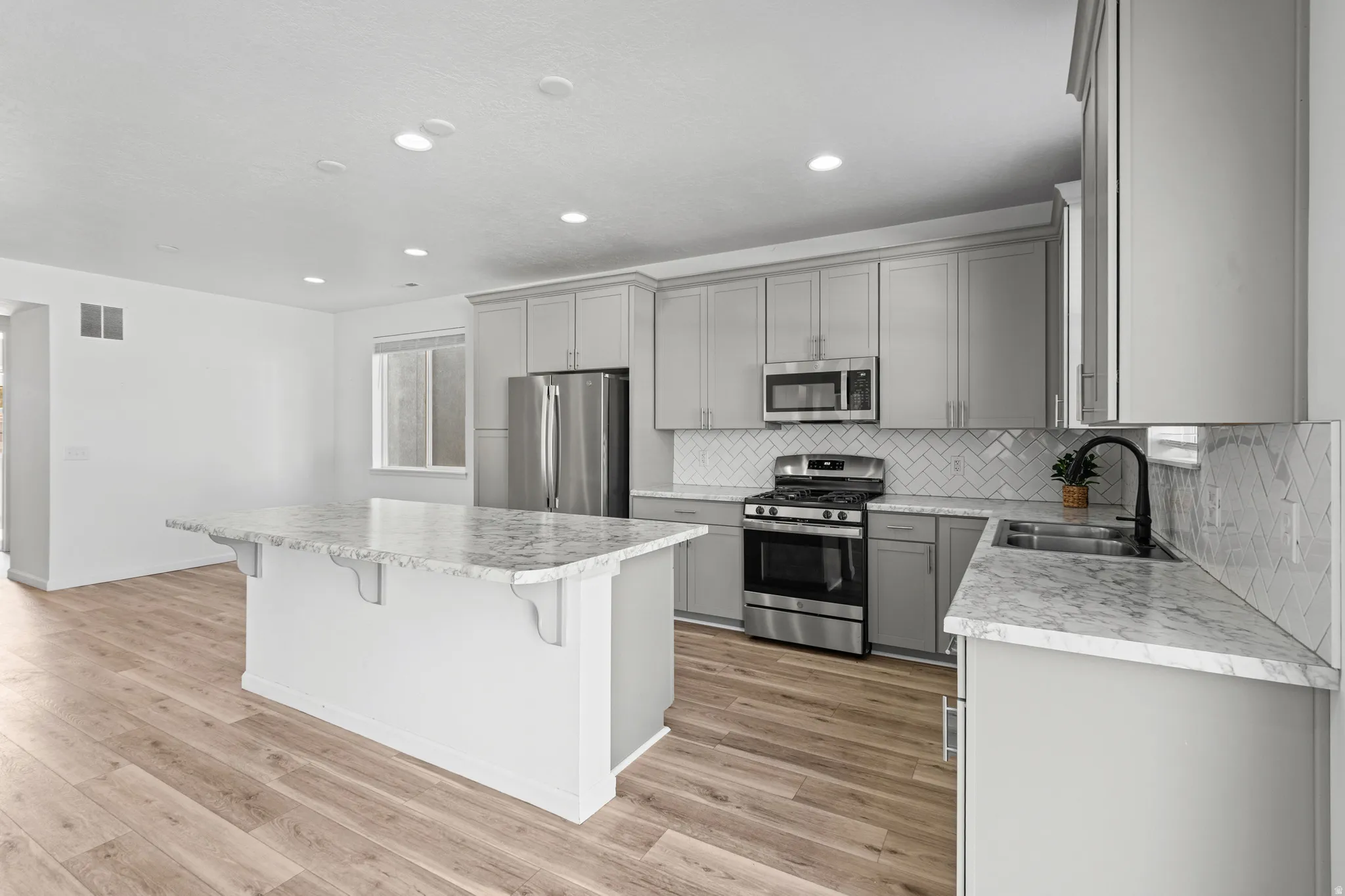 Contemporary kitchen featuring an expansive center island with stone countertops, stainless steel appliances, light gray cabinetry, a herringbone tile backsplash, and wood-finish flooring