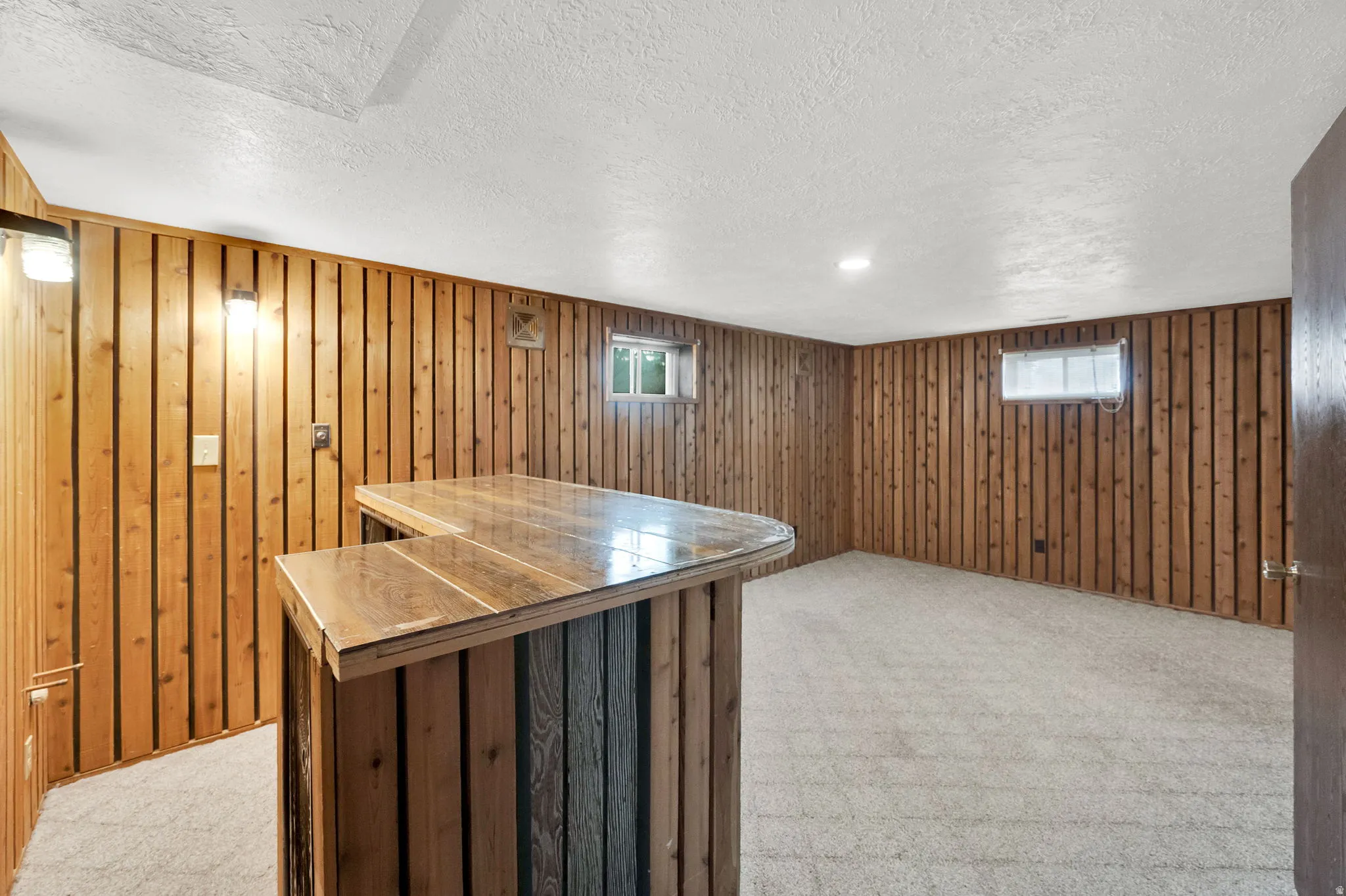 Basement with light colored carpet, a bar, plenty of natural light, wooden walls, and a textured ceiling