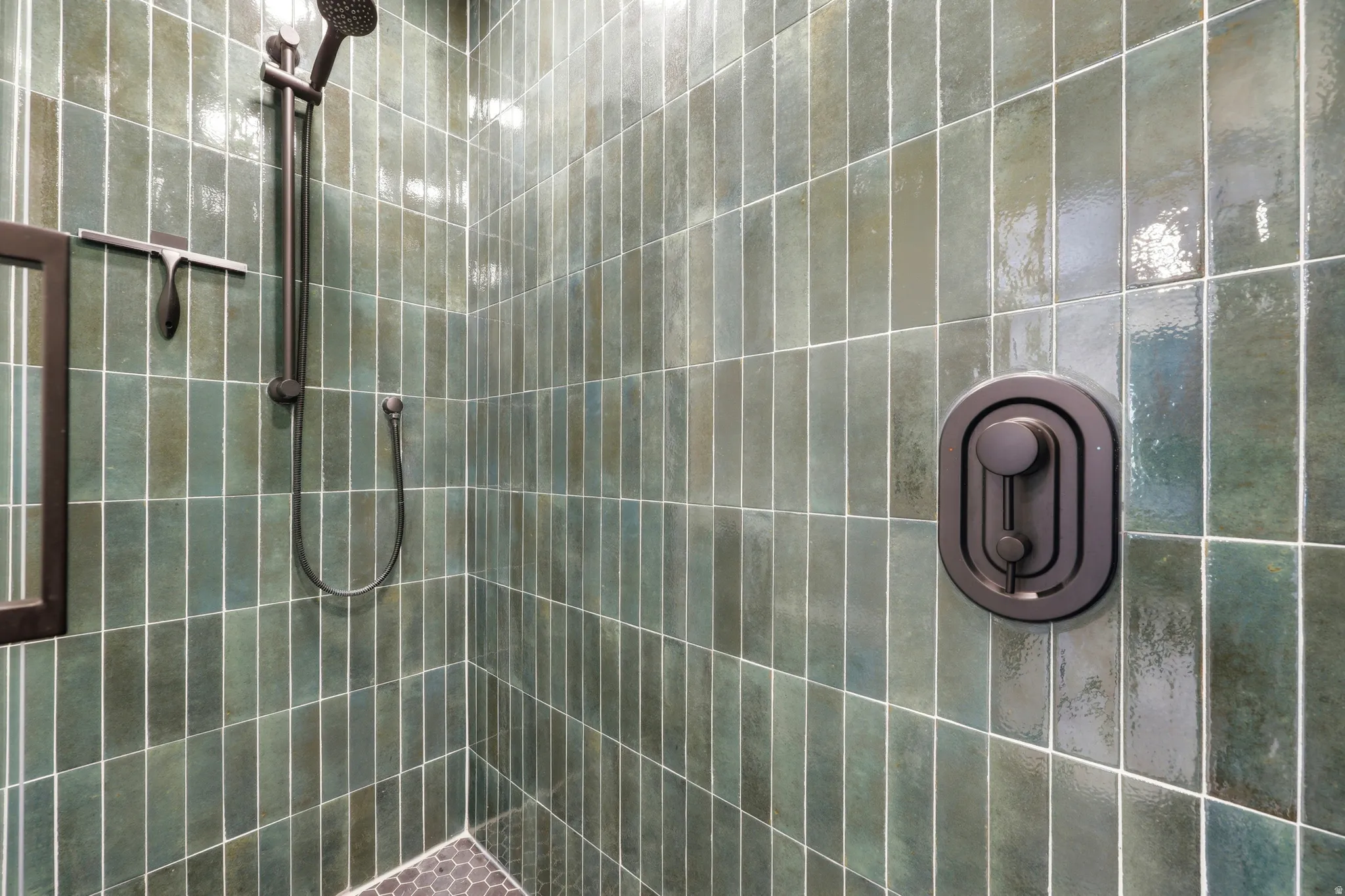 Shower stall featuring vertical green subway tile, matte black shower fixtures, a handheld showerhead, and a dark hexagonal tile floor