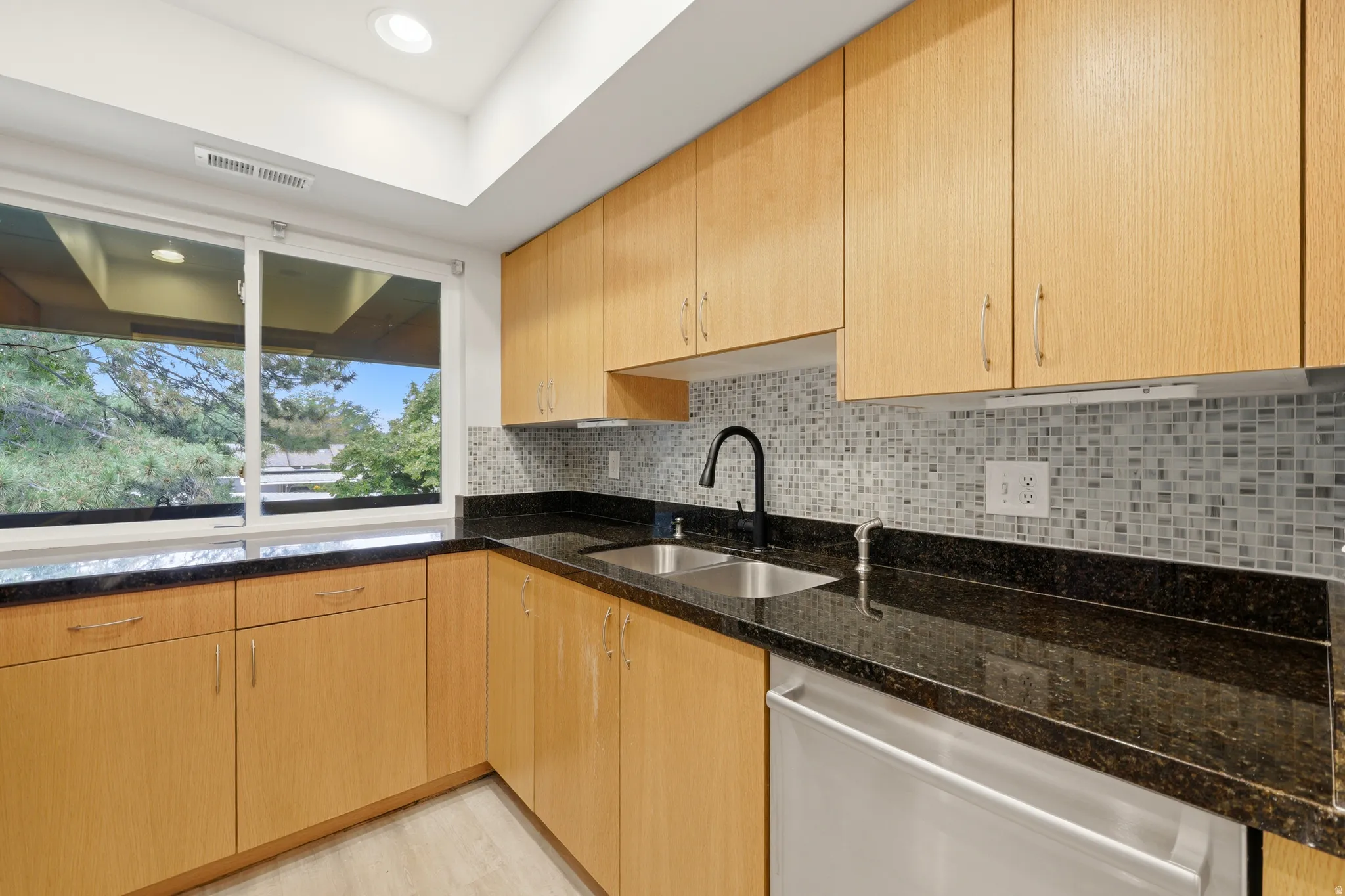 Kitchen featuring light wood cabinetry, dark stone countertops, a mosaic tile backsplash, stainless steel sink, and a black gooseneck faucet