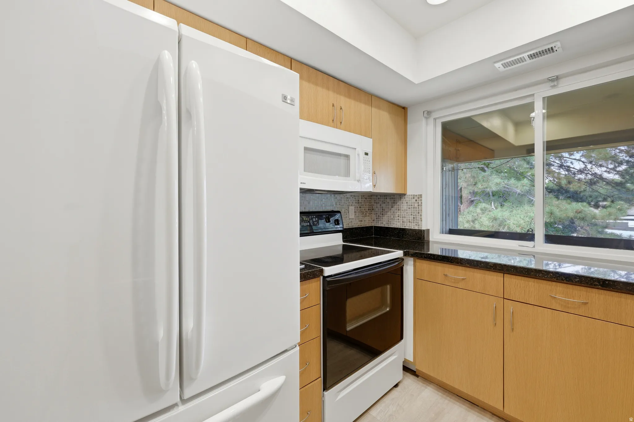 Kitchen featuring light wood cabinetry, dark countertops, white refrigerator, white oven, and white microwave