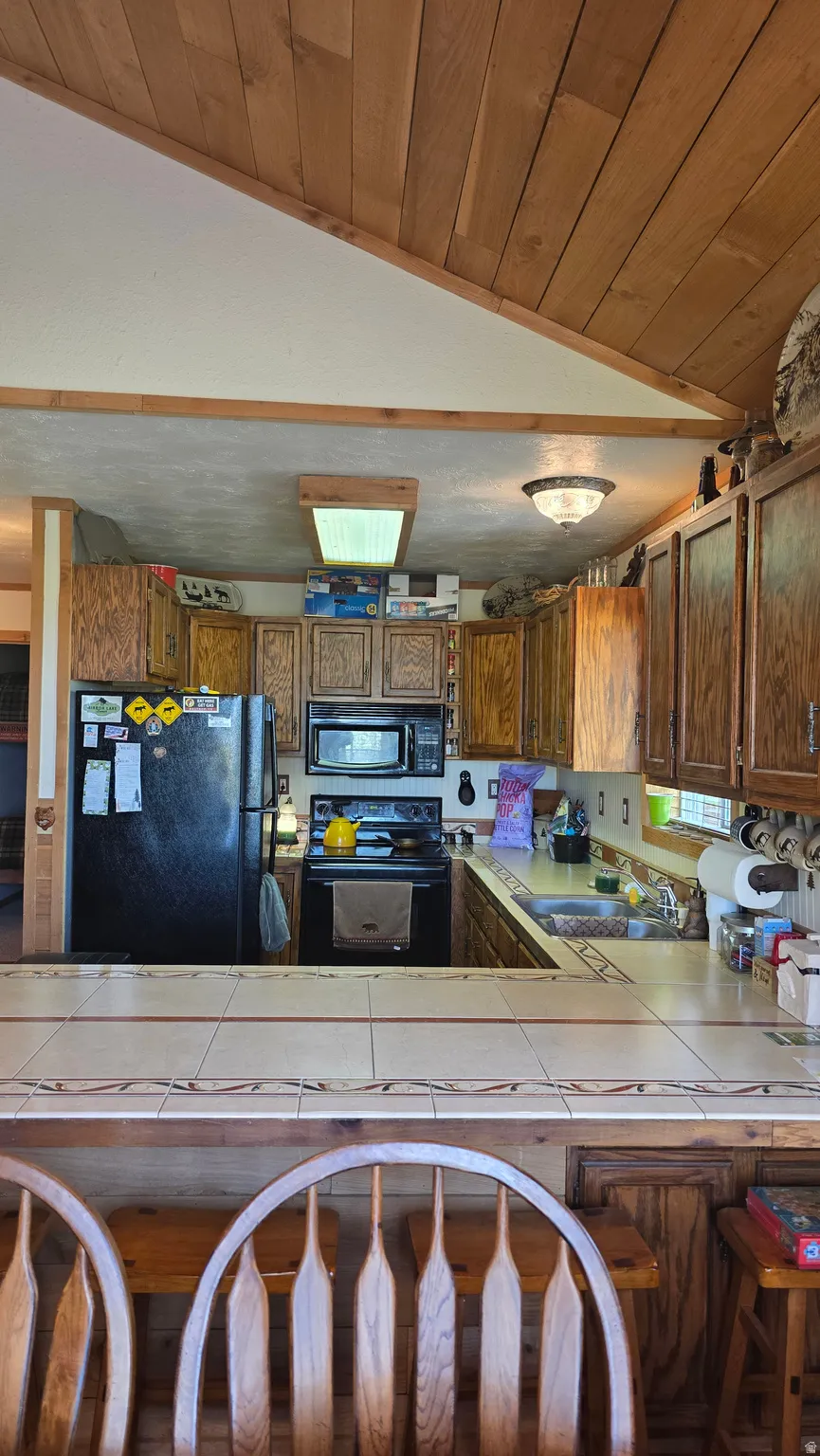 Kitchen with tile counters, a vaulted wood ceiling, wood finish cabinets, black appliances, and a peninsula