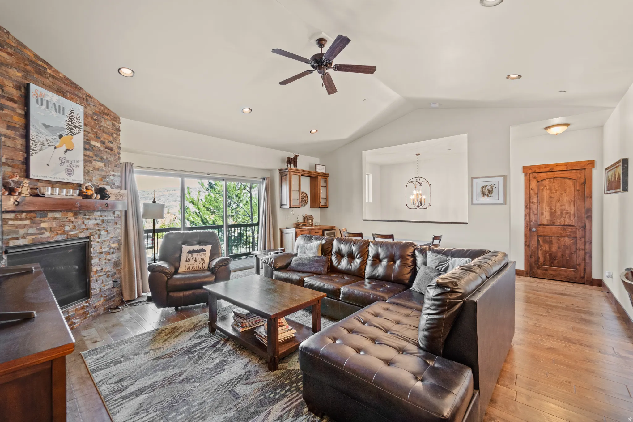 Living area featuring a ceiling fan, light wood finished floors, a chandelier, a stone fireplace, and vaulted ceiling