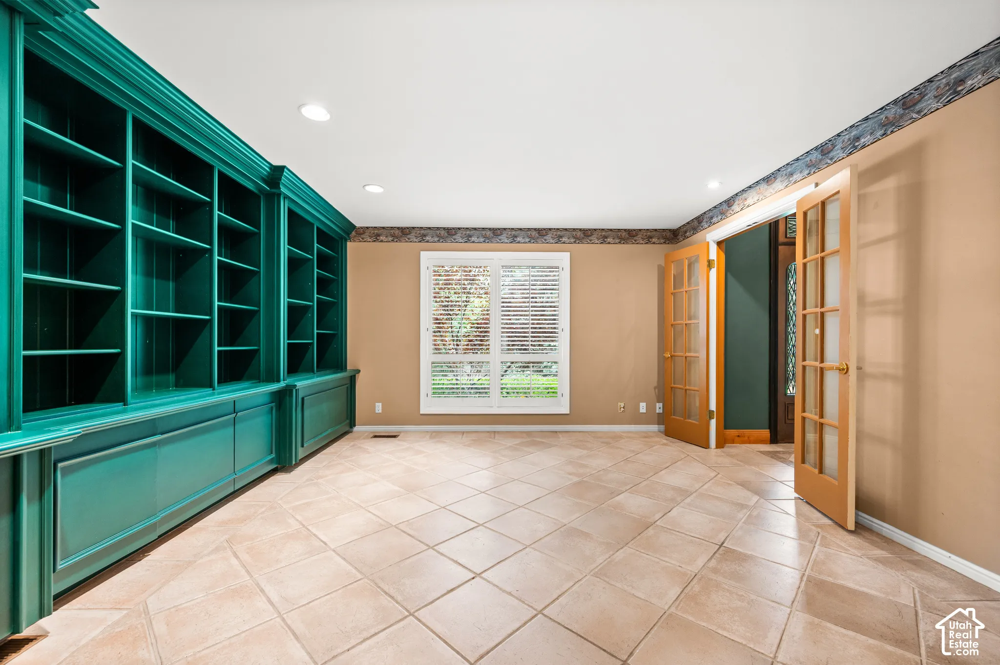 Expansive room featuring a wall of built-in shelving and cabinetry, light-toned tiled flooring, and a large window with plantation shutters.