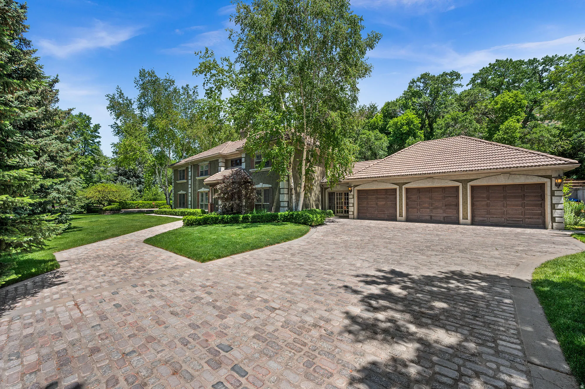 View of front of property with decorative driveway, 3-car garage, a tiled roof, a front yard, stone siding, and view of wooded area