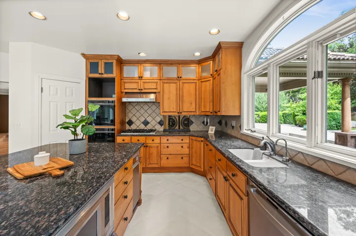 Kitchen featuring light wood cabinetry with glass-front accents, dark granite countertops, and a full suite of built-in appliances
