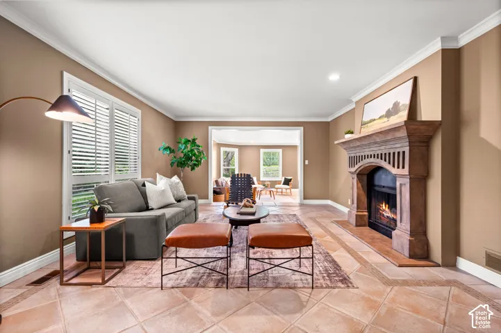 Spacious living area featuring a prominent fireplace with an ornate mantel, tile flooring with a decorative border, crown molding, white window shutters, and an arched doorway to an additional room