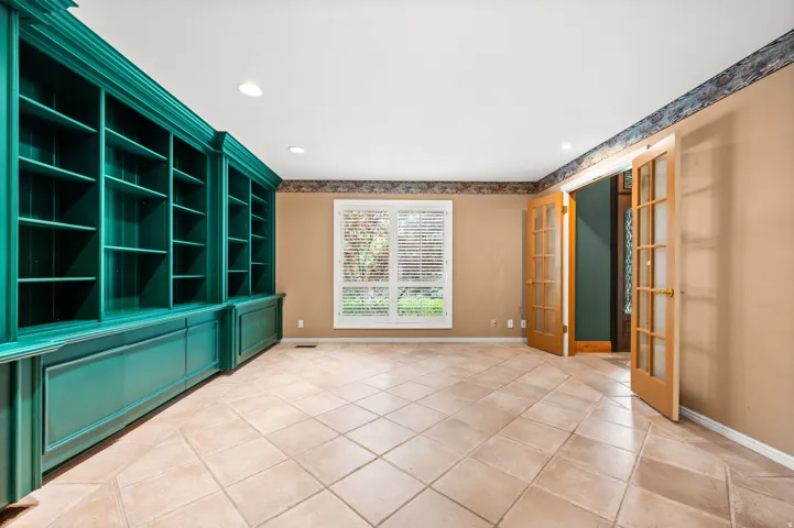 Expansive room featuring a wall of built-in shelving and cabinetry, light-toned tiled flooring, and a large window with plantation shutters.