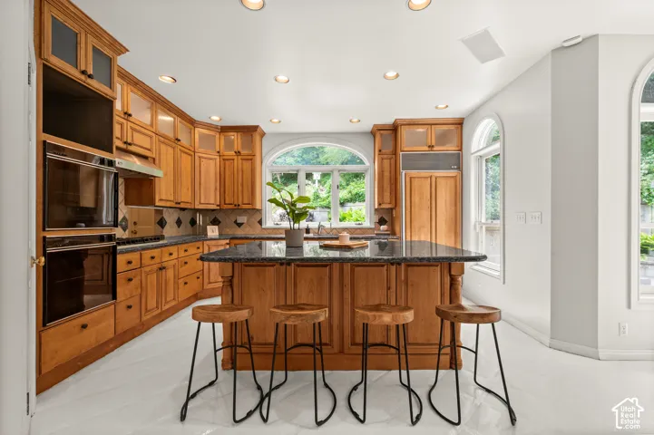 Kitchen featuring a central island with a dark countertop, light wood cabinetry, built-in double ovens, and arched windows