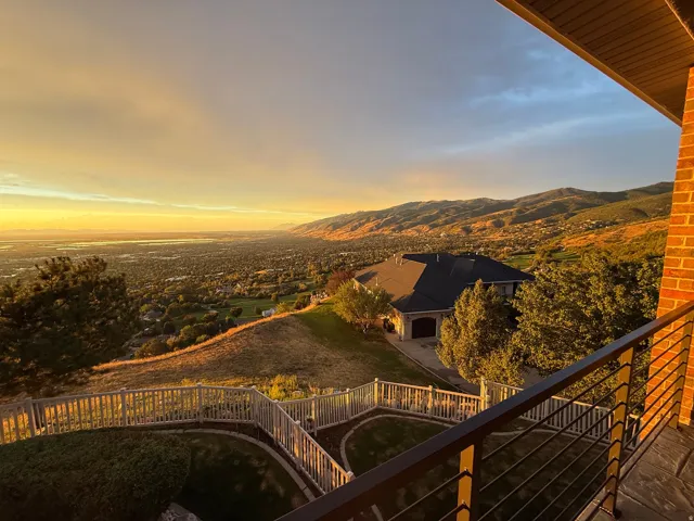 Expansive mountain and cityscape scenery, brick exterior, dark metal balcony railing, sloped landscaping with white fencing, and a dark shingle roof