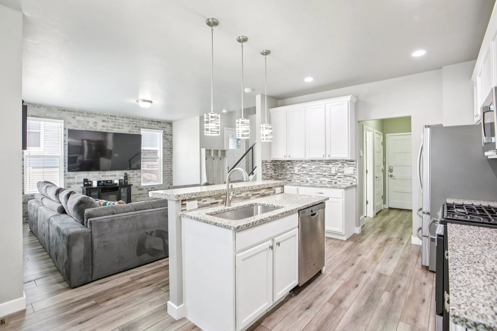 Kitchen featuring a kitchen island with sink, open floor plan, stainless steel appliances, white cabinetry, and brick wall